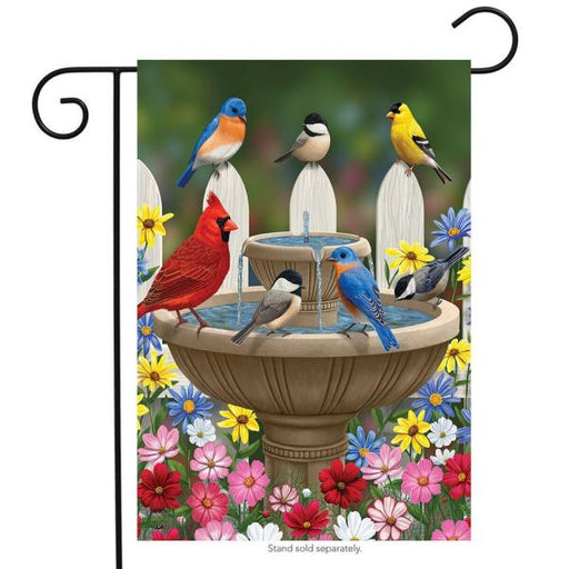 three chickadees, two bluebirds, a cardinal and a goldfinch gathered around a two tiered fountain surrounded by colorful flowers and a fence