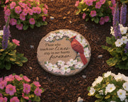 round, tan stepping stone with text that reads "those who touch our lives stay in our hearts forever" featuring a cardinal perched in a blossoming branch laying in garden bed. 