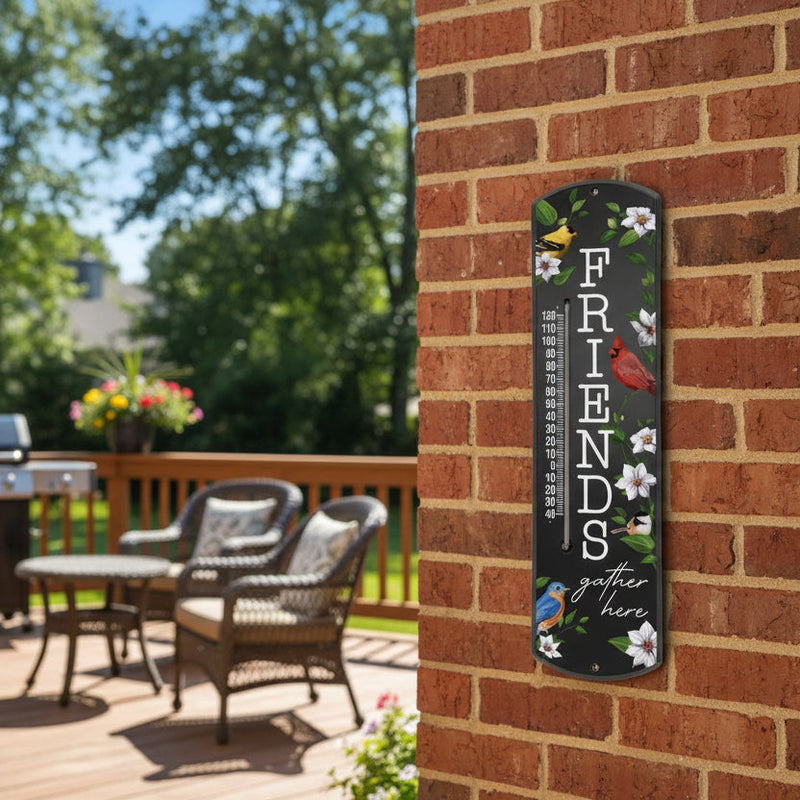 Friends gather here thermometer on wall outside with black background. featuring a goldfinch, cardinal, chickadee, and a bluebird surrounded by greenery and flowers 