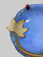 closer up view of ceramic bowl with blue glaze, moonseed leaf design, and small red ladybug on a beige background