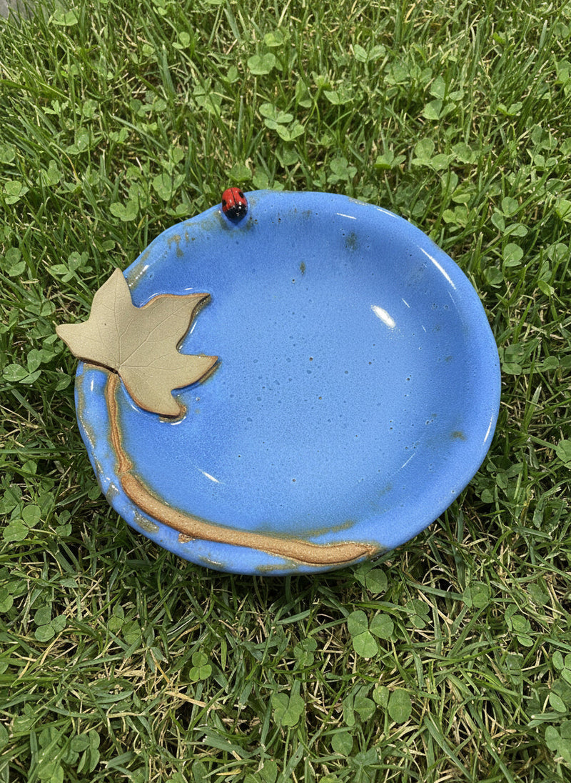 Ceramic bowl with blue glaze with a decorative moonseed vine leaf and a ladybug sitting in the grass