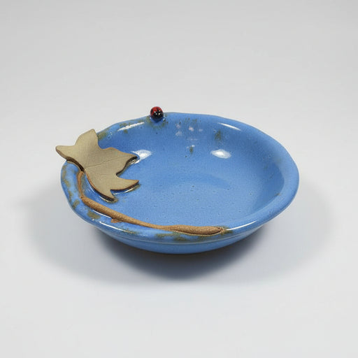 Ceramic bowl with blue glaze with a decorative moonseed vine leaf and a ladybug sitting in the grass