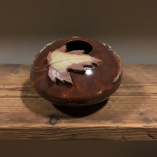 Brown ceramic pillow vase with a maple leaf on top, atop o a wooden shelf.