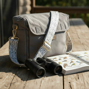 Gray shoulder bag with patterned strap on a wooden surface next to binoculars and a bird identification book.