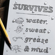 Sheet of paper with text 'SURVIVES water sweat grease & mud' and illustrations, with a pencil on a wooden surface.