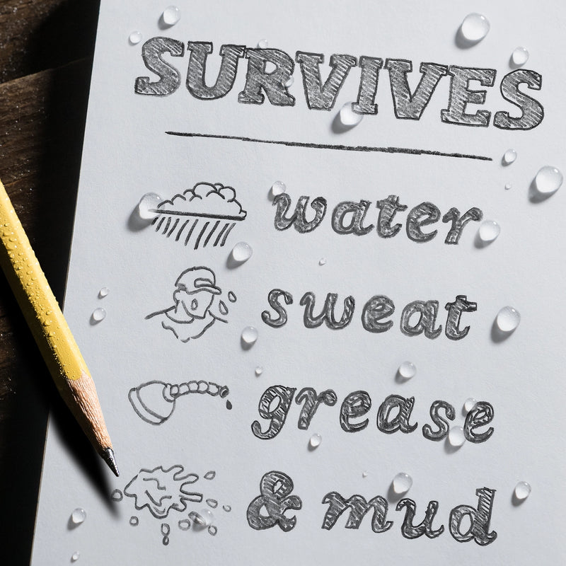 Sheet of paper with text 'SURVIVES water sweat grease & mud' and illustrations, with a pencil on a wooden surface.