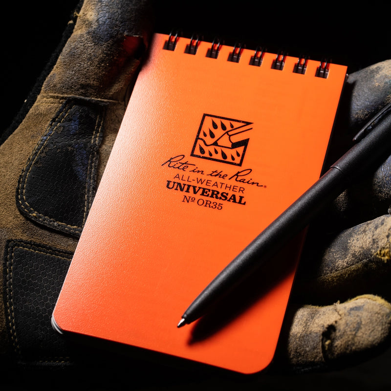 Orange notebook with 'Rite in the Rain' logo and a black pen being held in work gloves
