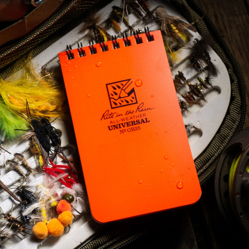 Orange notebook with 'Rite in the Rain' logo on a background of fishing equipment