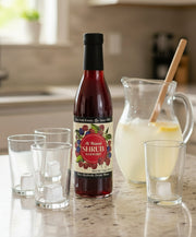 bottle of raspberry shrub next to empty glasses and a pitcher of lemonade