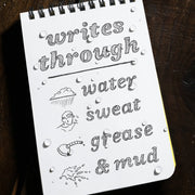 Notebook page with text 'writes through water, sweat, grease & mud' and illustrations on a wooden surface.
