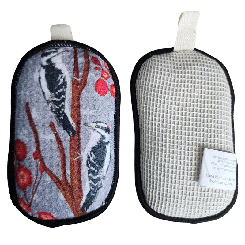 Two pouches sides of reversible reusable sponge, one with bird design and the other with cotton mesh textured surface, on a white background.