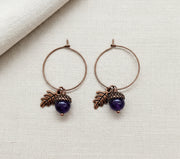 Pair of hoop earrings with leaf and purple arcorn charms on a white background