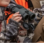 Person holding binoculars with camouflage clothing and gloves in the background