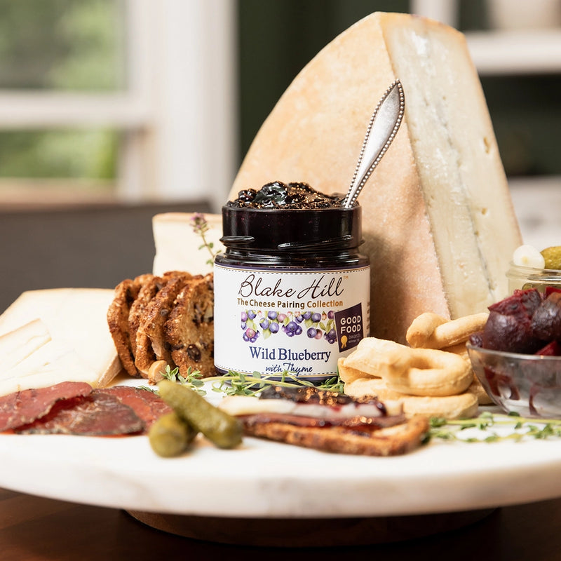 Platter with cheese, crackers, and a jar of Wild Blueberry with Thyme jam spread on a blurred background