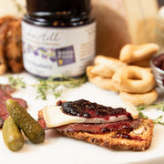 Platter with a slice of bread topped with cheese, meat, and Wild Blueberry with Thyme jam, accompanied by pickles and bagels.