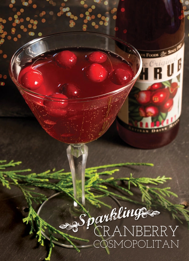 Cocktail with cranberries in a glass next to a bottle of cranberry juice on a dark surface.