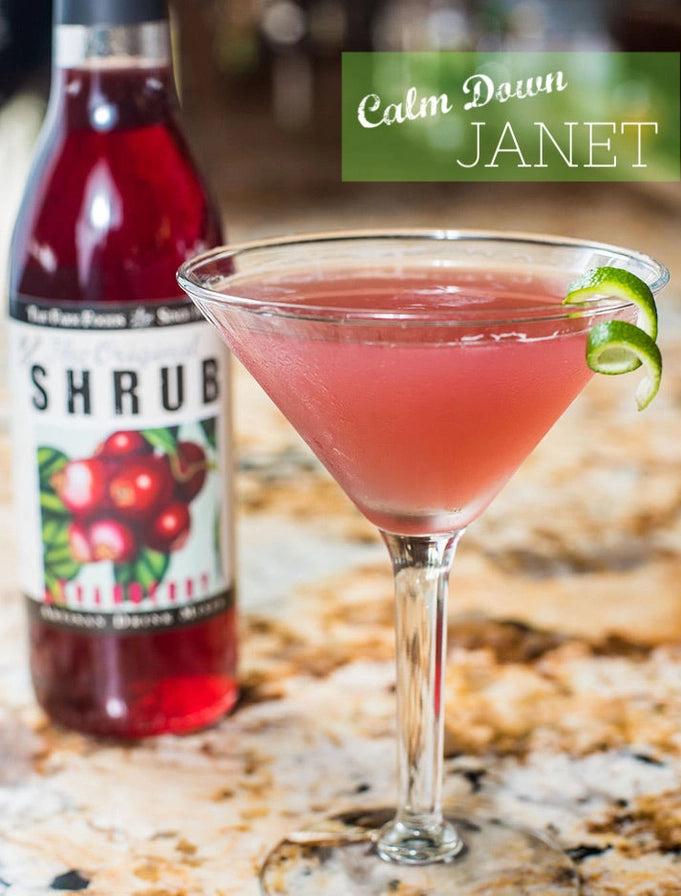 Pink cocktail in a martini glass with a bottle of 'Shrub' and a lime twist on a marble surface.
