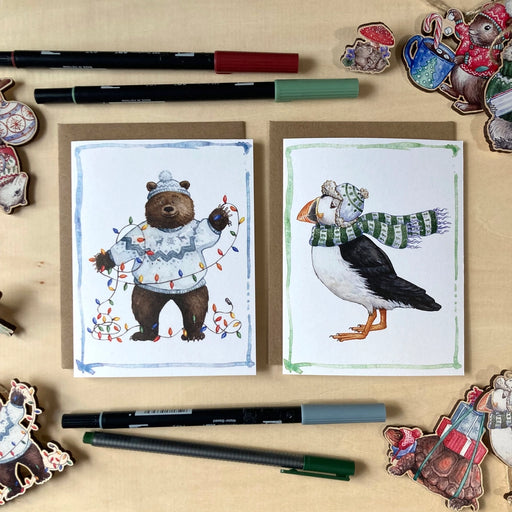 Two illustrated greeting cards on a wooden surface with markers and decorative items.