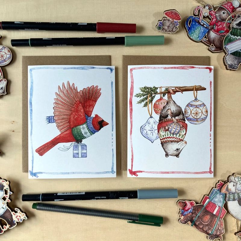 Two greeting cards with festive illustrations on a wooden surface with markers and decorative items.