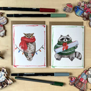 Two greeting cards with an owl and raccoon wearing scarves and a sweater on a wooden surface with pens and decorative items.