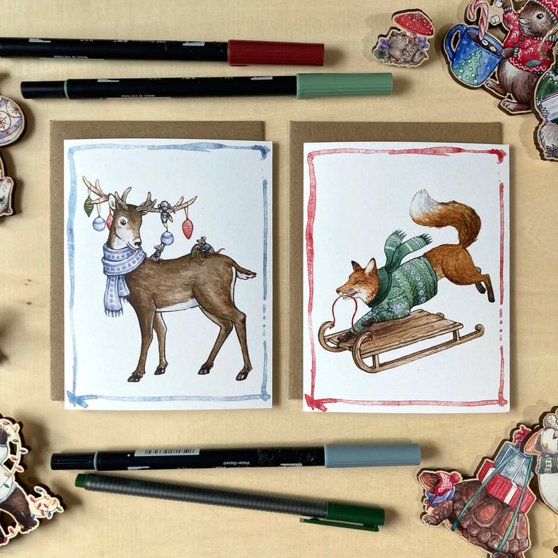 Two illustrated cards featuring a deer and a fox on a wooden surface with markers.