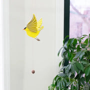 Yellow bird-shaped mobile against a white wall with a plant in the foreground