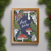 Card front 'Happy Holidays' text on a slate gray background, surrounded by ferns and mushrooms, in a brown box.