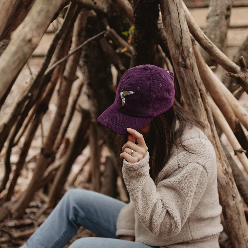 Person wearing a mulberry purple cap and beige jacket sitting against a wooden structure