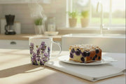 Cup of coffee with a floral design and a piece of blueberry cake on a plate in a kitchen setting.