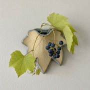 A stoneware grapevine-shaped wall vase filled with a sprig of moonseed vine against a neutral off-white wall.