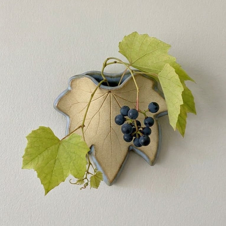 A stoneware grapevine-shaped wall vase filled with a sprig of moonseed vine against a neutral off-white wall.