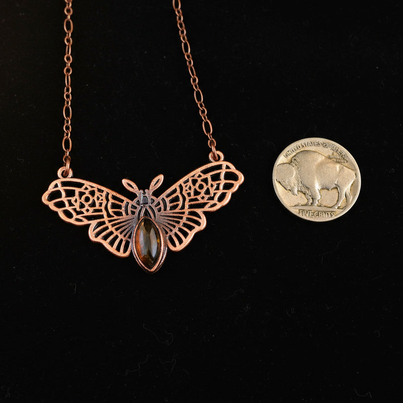 Copper witch moth necklace next to a quarter on a dark background