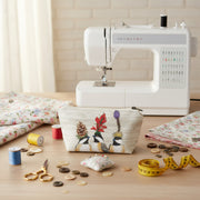 Canvas bag on a wooden table with fabric, buttons, scissors, and a measuring tape with a sewing machine in the background