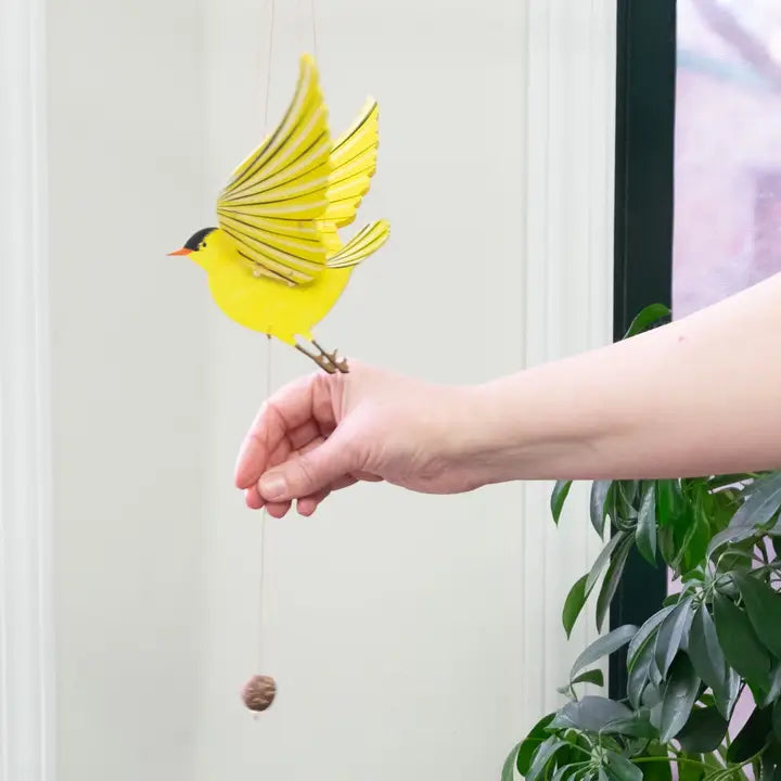 Hand pulling the pull string on a yellow bird-shaped mobile against a white wall with a plant in the corner.